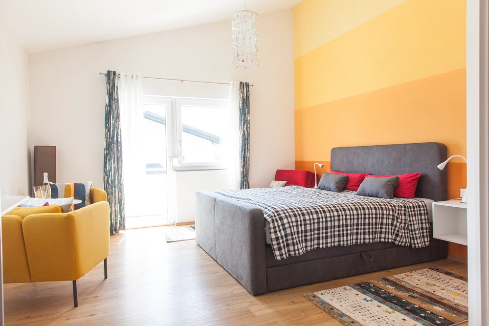 „Hahns Herberge“ – Interiorfoto; Seitenansicht Schlafzimmer; Die Farben sind in verschiedenen Orangetönen und rot gehalten, aufgeräumtes, helles Ambiente, Blick durchs Fenster.